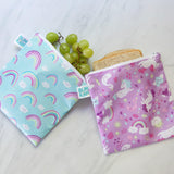 Bumkins Reusable Snack Bag, Large 2-Pack: Rainbows & Unicorns holds grapes and a sandwich on a marble surface.
