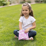 A girl with long hair sits on grass, smiling while holding a Bumkins Reusable Snack Bag, Large 2-Pack: Rainbows & Unicorns.