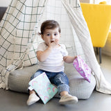A toddler covers their mouth, sitting in a checkered fabric tent and holding Bumkins Reusable Snack Bag, Large 2-Pack: Rainbows & Unicorns.