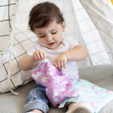 A toddler in jeans opens a Bumkins Reusable Snack Bag, Large 2-Pack: Rainbows & Unicorns while sitting inside a cloth tent.