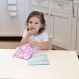 A young girl in a white shirt enjoys snacks from Bumkins Reusable Snack Bag, Large 2-Pack: Rainbows & Unicorns at a white table.