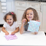 Two smiling children hold Bumkins Reusable Snack Bag, Large 2-Pack: Rainbows & Unicorns at a white table in a kitchen.
