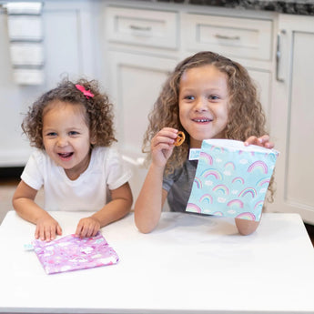 Two smiling children hold Bumkins Reusable Snack Bag, Large 2-Pack: Rainbows & Unicorns at a white table in a kitchen.