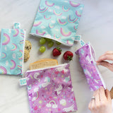 A person zips Bumkins Reusable Snack Bag, Small 2-Pack: Rainbows & Unicorns on a marble surface with other bags and snacks close by.