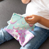 Close-up of a child on a gray couch holding a Bumkins Reusable Snack Bag, Small 2-Pack: Rainbows & Unicorns with another on their lap.