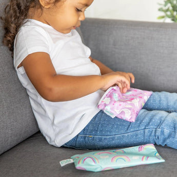 A child in jeans and a white shirt sits with Bumkins Reusable Snack Bag, Small 2-Pack: Rainbows & Unicorns on a gray couch.