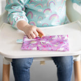 Close-up of a child in a rainbow bib with a purple Bumkins Reusable Snack Bag, Small 2-Pack: Rainbows & Unicorns on a high chair tray.