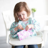 A toddler in a high chair wearing a blue bib enjoys snacks from a Bumkins Reusable Snack Bag, Small 2-Pack: Rainbows & Unicorns.