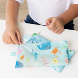 Close-up of a child zipping a Bumkins Reusable Snack Bag, Small 2-Pack: Ocean Life & Whale Tail on top of another bag on a white table.