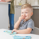A child smiles while snacking from Bumkins Reusable Snack Bag, Small 2-Pack: Ocean Life & Whale Tail; an adult stands in the background.