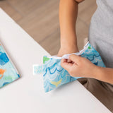 Close-up of a child reaching into a Bumkins Reusable Snack Bag, Small 2-Pack: Ocean Life & Whale Tail at a white table.