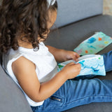 A curly-haired child, in a white top and blue jeans, holds a Bumkins Reusable Snack Bag, Small 2-Pack: Ocean Life & Whale Tail on a couch.