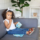 A toddler with curly hair and a bow enjoys snacks from Bumkins Reusable Snack Bag, Small 2-Pack: Ocean Life & Whale Tail on a gray sofa.