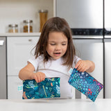 A young girl holds Bumkins Reusable Snack Bag, Small 2-Pack: Jungle & Animal Prints in a bright kitchen.