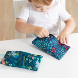 A child in a white shirt examines Bumkins Reusable Snack Bag, Small 2-Pack: Jungle & Animal Prints on a white table.