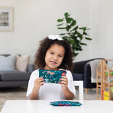 A curly-haired child holds a Bumkins Reusable Snack Bag, Small 2-Pack: Jungle & Animal Prints with another bag resting on a white table.