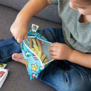 A child on a gray couch opens a blue Bumkins Reusable Snack Bag, Small 2-Pack: Super Mario™, revealing veggie sticks inside.