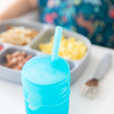Close-up of Bumkins Silicone Straw Cup with Lid: Blue on a white table, with a gray plate of food and fork blurred in the background.