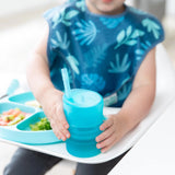 A toddler in a blue bib holds a Bumkins Silicone Straw Cup with Lid: Blue, sitting in a high chair beside a plate of varied foods.