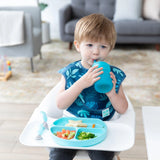 A child in a high chair wearing a blue bib drinks from a Bumkins Silicone Straw Cup with Lid: Blue, with a blue plate of food nearby.