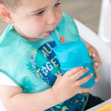 A toddler uses a Bumkins Silicone Straw Cup with Lid: Blue, drinking independently indoors while wearing a bib.