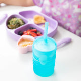 Bumkins Silicone Straw Cup with Lid: Blue sits on a white table in front of a purple sectioned plate of food.
