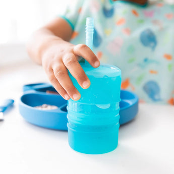A toddler holds a Bumkins Silicone Straw Cup with Lid: Blue, while a silicone plate sits on the white table in the background.
