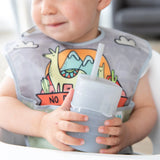 A toddler with a colorful dinosaur bib holds a Bumkins Silicone Straw Cup with Lid: Gray.