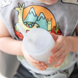 A child proudly holds a Bumkins Silicone Straw Cup with Lid: Gray while wearing a dinosaur bib.