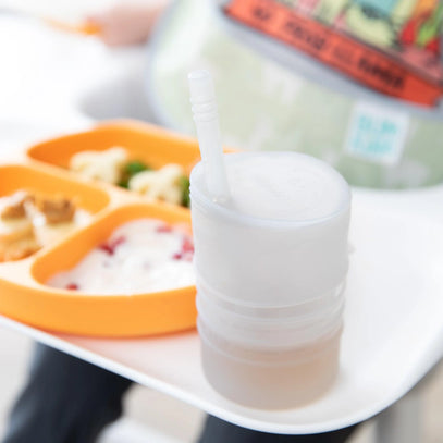 Close-up of a white cup with a lid and straw next to an orange divided plate holding snacks. The plate has compartments with food, visible on a white surface, and a person seated in the background.