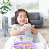 A toddler with a bow holds a Bumkins Silicone Straw Cup with Lid: Gray in a high chair next to a purple plate.