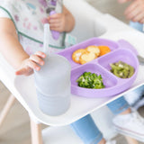 A child in a high chair reaches for a Bumkins Silicone Straw Cup with Lid: Gray next to a lavender plate.