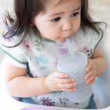 A child uses a Bumkins Silicone Straw Cup with Lid: Gray, honing their drinking skills while wearing a floral bib.