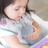 A toddler with a floral bib sits at a table, holding a Bumkins Silicone Straw Cup with Lid: Gray, exploring independent drinking.