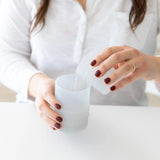 In a white shirt, a person uses a cup to pour water into a Bumkins Silicone Straw Cup with Lid: Gray at a white table.