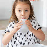 A young girl with brown hair, in a heart-patterned bib, sips from a Bumkins Silicone Straw Cup with Lid: Gray.