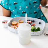 A child in a blue bib sits at a white table with a Bumkins Silicone Straw Cup with Lid: Gray and a gray plate of food.