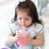 A toddler in a floral bib sips from a Bumkins Silicone Straw Cup with Lid: Pink, with a bow in her hair.