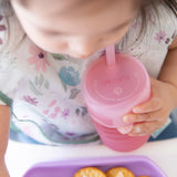 A toddler confidently drinks from a Bumkins Silicone Straw Cup with Lid: Pink, while gazing at a purple tray of crackers.