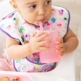 A baby with a colorful bib sips from a Bumkins Silicone Straw Cup with Lid: Pink in a high chair.