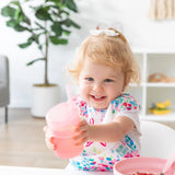 A smiling toddler in a high chair holds a Bumkins Silicone Straw Cup with Lid: Pink, with a plant and shelves in the background.