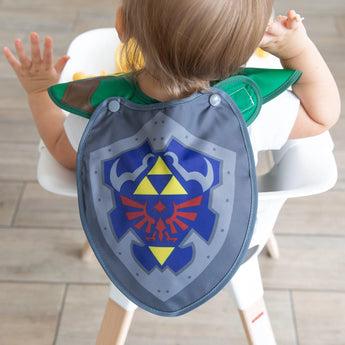 A toddler in a high chair faces away from the camera, showcasing the shield-shaped cape of Bumkins Caped SuperBib®: The Legend of Zelda™ Link.