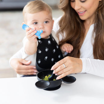 A baby with in a bib holds a Bumkins Silicone Dipping Spoons: Mickey Mouse (Black and Blue), while an adult feeds them from a bowl.