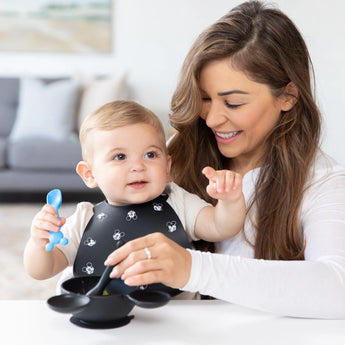 A baby in a bib sits on a woman's lap as she feeds them with a Bumkins Silicone Dipping Spoons: Mickey Mouse (Black and Blue) from a bowl.