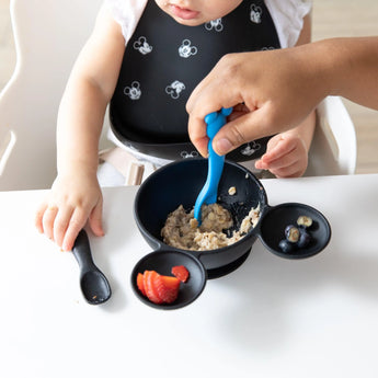 A baby in a high chair in a bib grabs a Bumkins Silicone Dipping Spoons: Mickey Mouse (Black and Blue), while an adult feeds them from a bowl.