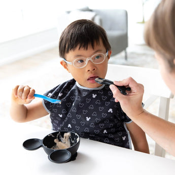 A child with glasses self-feeds from a bowl using a Bumkins Silicone Dipping Spoons: Mickey Mouse (Black and Blue), assisted by an adult.