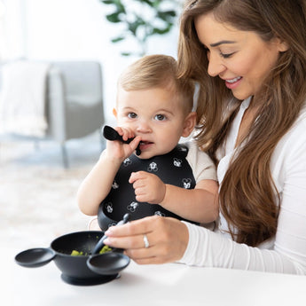 A woman smiles as a baby chews a Bumkins Silicone Dipping Spoons: Mickey Mouse (Classic Black), with a black bowl of food on a table.