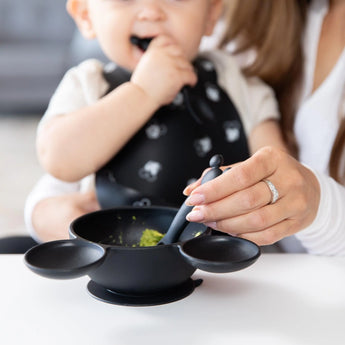 A baby in a black bib is fed green food with a Bumkins Silicone Dipping Spoons: Mickey Mouse (Classic Black) from a black bowl by an adult.