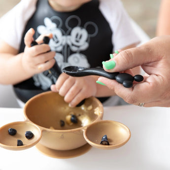 An adult helps feed a child from a gold bowl of blueberries with a Bumkins Silicone Dipping Spoons: Mickey Mouse (Classic Black).