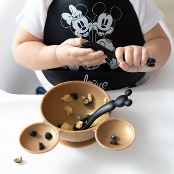 A child in a black bib holds a Bumkins Silicone Dipping Spoons: Mickey Mouse (Classic Black), with a gold bowl of food on a high chair tray.